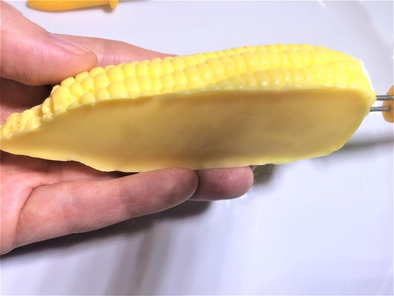 以下が含まれることがあります： 黄色いトウモロコシの穂が、半分に縦に切られて手に持たれています。穂は薄い黄色で、滑らかでワックスのような質感です。