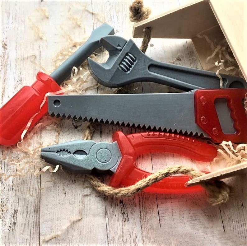 Op de afbeelding: Een set van vier speelgoedgereedschappen, waaronder een moersleutel, een zaag, een schroevendraaier en een tang. De gereedschappen zijn gemaakt van plastic en hebben rode en grijze handvatten.