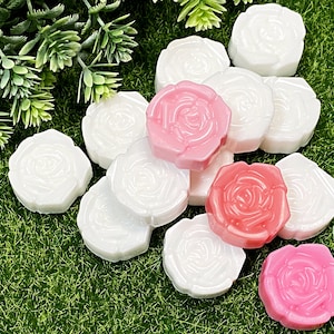 May include: Rose-shaped soaps in white and pink, arranged on a green surface. The soaps have a detailed floral design, with some greenery in the background. The soaps are likely for decorative or cosmetic use.