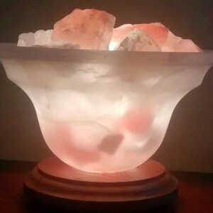 K&ouml;nnte beinhalten: Eine wei&szlig;e Salzlampe mit einem Holzsockel und rosa Himalaya-Salzkristallen im Inneren. Die Lampe ist eingeschaltet und leuchtet.