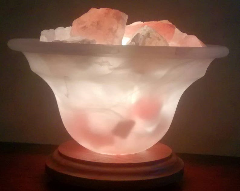 K&ouml;nnte beinhalten: Eine wei&szlig;e Salzlampe mit einem Holzsockel und rosa Himalaya-Salzkristallen im Inneren. Die Lampe ist eingeschaltet und leuchtet.