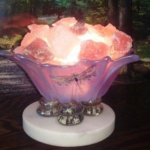 May include: A pink Himalayan salt lamp with a dragonfly design on the base. The lamp is lit and the salt crystals are glowing. The lamp is sitting on a white marble base.