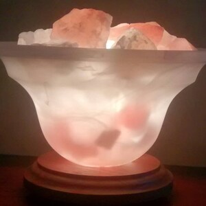 K&ouml;nnte beinhalten: Eine wei&szlig;e Salzlampe mit einem Holzsockel. Die Lampe ist mit rosa Himalaya-Salzkristallen gef&uuml;llt und von innen beleuchtet, wodurch ein warmes Leuchten entsteht.
