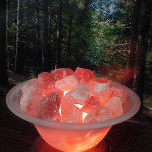 May include: A Himalayan salt lamp with a frosted glass bowl filled with pink Himalayan salt crystals. The lamp is on a wooden base and is glowing orange.