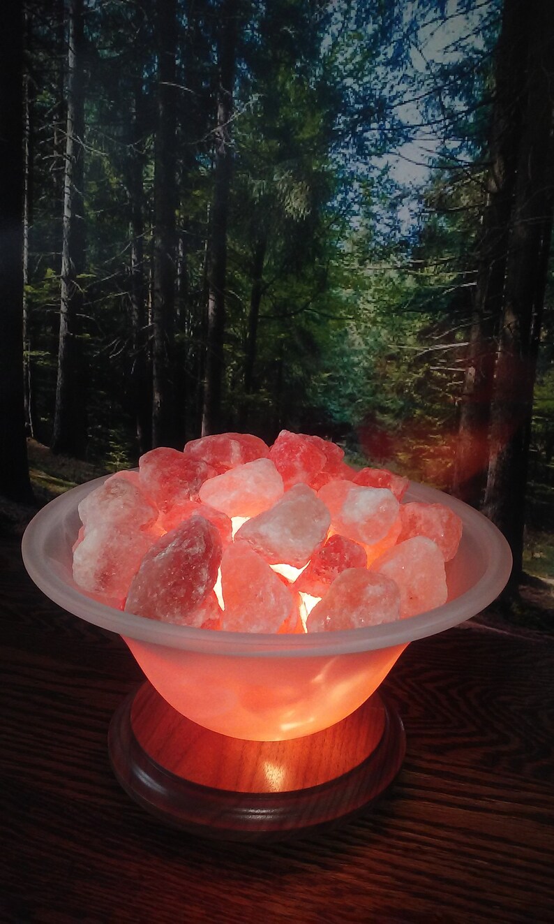 May include: A Himalayan salt lamp with a frosted glass bowl filled with pink Himalayan salt crystals. The lamp is on a wooden base and is glowing orange.