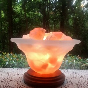 K&ouml;nnte beinhalten: Eine Himalaya-Salzlampe in Form einer Schale mit einem Holzsockel. Die Salzlampe ist eingeschaltet und leuchtet orange.