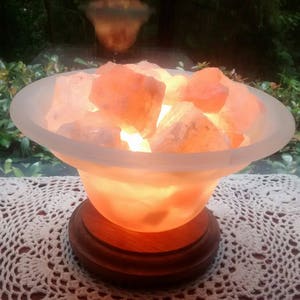 K&ouml;nnte beinhalten: Eine Himalaya-Salzlampe mit einer mattierten Glasschale, gef&uuml;llt mit rosa Salzkristallen. Die Lampe ist eingeschaltet und leuchtet, wobei sie ein warmes orangefarbenes Licht abgibt. Die Lampe steht auf einem Holzsockel und befindet sich auf einer wei&szlig;en H&auml;keldecke.