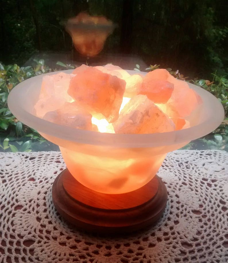 K&ouml;nnte beinhalten: Eine Himalaya-Salzlampe mit einer mattierten Glasschale, gef&uuml;llt mit rosa Salzkristallen. Die Lampe ist eingeschaltet und leuchtet, wobei sie ein warmes orangefarbenes Licht abgibt. Die Lampe steht auf einem Holzsockel und befindet sich auf einer wei&szlig;en H&auml;keldecke.