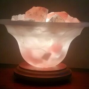 K&ouml;nnte beinhalten: Eine wei&szlig;e Salzlampe mit einem Holzsockel. Die Lampe ist mit rosa Himalaya-Salzkristallen gef&uuml;llt und von innen beleuchtet, wodurch ein warmes Leuchten entsteht.