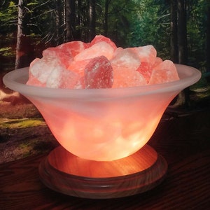 May include: A Himalayan salt lamp with a frosted glass bowl filled with pink Himalayan salt crystals. The lamp is on a wooden base and is lit, casting a warm glow.