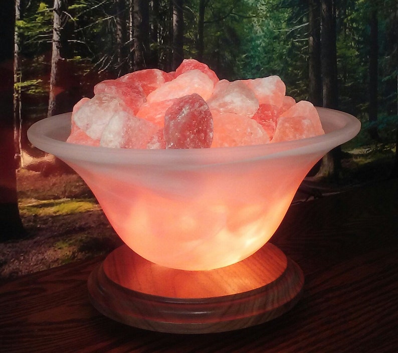 May include: A Himalayan salt lamp with a frosted glass bowl filled with pink Himalayan salt crystals. The lamp is on a wooden base and is lit, casting a warm glow.