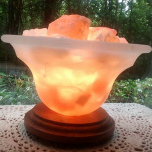 K&ouml;nnte beinhalten: Eine Himalaya-Salzlampe in Form einer Schale mit einem Holzsockel. Die Salzlampe ist eingeschaltet und leuchtet orange.