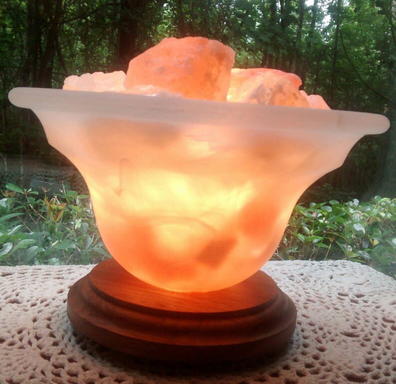 K&ouml;nnte beinhalten: Eine Himalaya-Salzlampe in Form einer Schale mit einem Holzsockel. Die Salzlampe ist eingeschaltet und leuchtet orange.