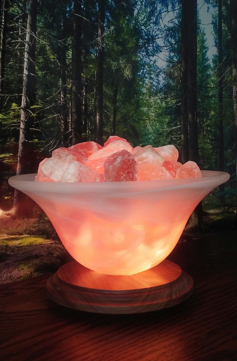 May include: A Himalayan salt lamp with a white frosted glass bowl filled with pink Himalayan salt crystals. The lamp is on a wooden base and is lit, casting a warm glow.