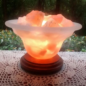 K&ouml;nnte beinhalten: Eine Himalaya-Salzlampe mit einer mattierten Glasschale, gef&uuml;llt mit rosa Salzkristallen. Die Lampe steht auf einem Holzsockel und ist eingeschaltet, wodurch ein warmes Licht entsteht.