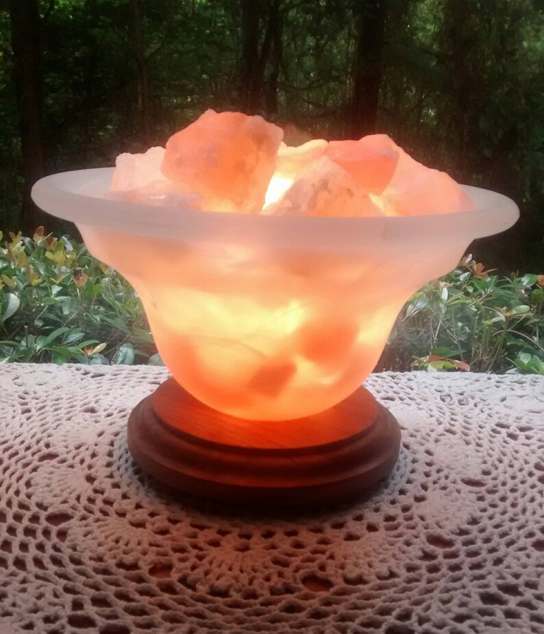 K&ouml;nnte beinhalten: Eine Himalaya-Salzlampe mit einer mattierten Glasschale, gef&uuml;llt mit rosa Salzkristallen. Die Lampe steht auf einem Holzsockel und ist eingeschaltet, wodurch ein warmes Licht entsteht.