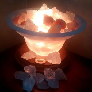 May include: A Himalayan salt lamp with a white ceramic base and a wooden base. The lamp is lit and the salt crystals are glowing orange.
