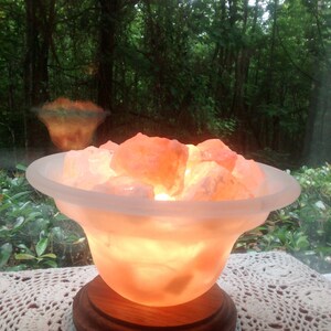 K&ouml;nnte beinhalten: Eine Himalaya-Salzlampe mit einer mattierten Glasschale, gef&uuml;llt mit rosa Himalaya-Salzkristallen. Die Lampe ist eingeschaltet und leuchtet in einem warmen Orangeton. Die Lampe steht auf einem Holzsockel und befindet sich auf einer wei&szlig;en H&auml;keldeckchen.