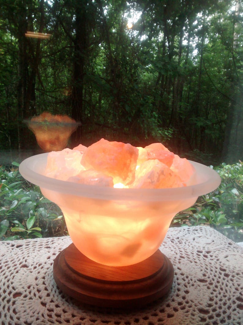 K&ouml;nnte beinhalten: Eine Himalaya-Salzlampe mit einer mattierten Glasschale, gef&uuml;llt mit rosa Himalaya-Salzkristallen. Die Lampe ist eingeschaltet und leuchtet in einem warmen Orangeton. Die Lampe steht auf einem Holzsockel und befindet sich auf einer wei&szlig;en H&auml;keldeckchen.