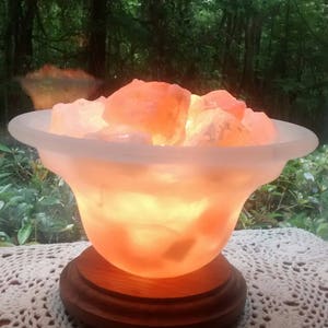 K&ouml;nnte beinhalten: Eine Himalaya-Salzlampe mit einer mattierten Glasschale, gef&uuml;llt mit rosa Salzkristallen. Die Lampe steht auf einem Holzsockel und ist eingeschaltet, wodurch ein warmes Licht entsteht.