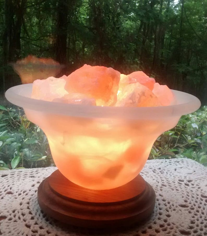 K&ouml;nnte beinhalten: Eine Himalaya-Salzlampe mit einer mattierten Glasschale, gef&uuml;llt mit rosa Salzkristallen. Die Lampe steht auf einem Holzsockel und ist eingeschaltet, wodurch ein warmes Licht entsteht.