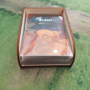 K&ouml;nnte beinhalten: Eine braune Holzkiste mit einem transparenten Kunststoffeinsatz enth&auml;lt einen Stapel Karten. Die oberste Karte zeigt ein Zombiebild und den aufgedruckten Text "EVENT".