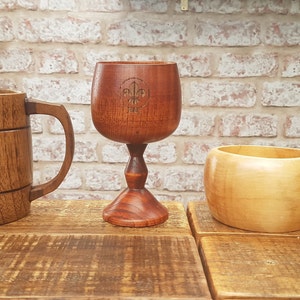 Może przedstawiać: Trzy drewniane kubki do picia, jeden w kształcie kielicha z motywem lilii, jeden wysoki i cylindryczny z motywem lilii, a jeden zaokrąglony z dwoma otworami w uchwycie.