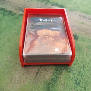 K&ouml;nnte beinhalten: Ein roter Plastikkartenhalter mit einem Stapel Karten darin. Die oberste Karte tr&auml;gt den Aufdruck "EVENT". Die Karten zeigen eine Zombie-Illustration.