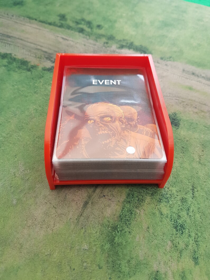 K&ouml;nnte beinhalten: Ein roter Plastikkartenhalter mit einem Stapel Karten darin. Die oberste Karte tr&auml;gt den Aufdruck "EVENT". Die Karten zeigen eine Zombie-Illustration.