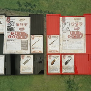 Puede incluir: Dos bandejas de plástico rojo y negro con compartimentos para guardar cartas y fichas de juego. Las bandejas están etiquetadas como "REGAN" y "MARTINEZ" y presentan retratos de personajes, estadísticas y reglas especiales. Las cartas incluyen "Sombrero de la Suerte", "Extintor", "Radio", "Herramienta de Demolición", "Fusil de Asalto M16" y "Amuleto de la Suerte".
