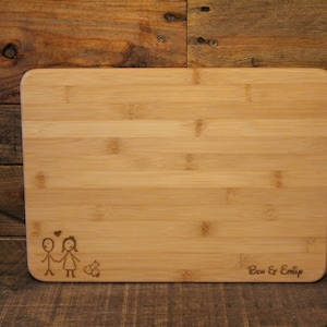 Puede incluir: Una tabla de cortar de bambú natural con una ilustración dibujada a mano de una pareja y un gato. La tabla está grabada con el texto "Ben & Emily".