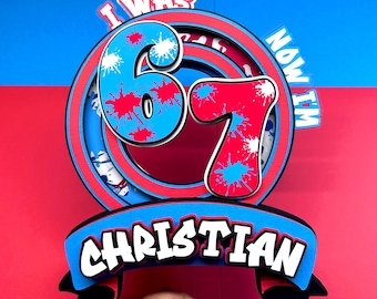 May include: A layered birthday cake topper with the text "I WAS 67 NOW I'M" and the name "CHRISTIAN". The numbers 6 and 7 are blue and red with paint splatter details. The background is red and blue.