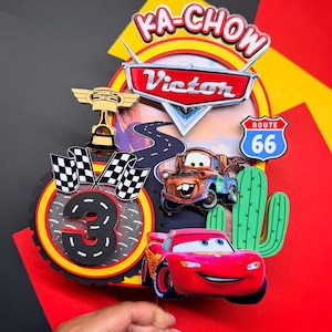 Peut inclure: Décoration de gâteau sur le thème de Cars, avec Flash McQueen, Martin et un panneau Route 66. Le design comprend un drapeau à damier, un trophée et le chiffre 3. Les textes "KA-CHOW" et "Victor" sont également visibles.