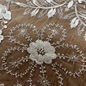 May include: White floral lace fabric with pearl and sequin embellishments. The fabric features a delicate floral pattern with intricate details. The lace is soft and flowing, making it ideal for a variety of projects.