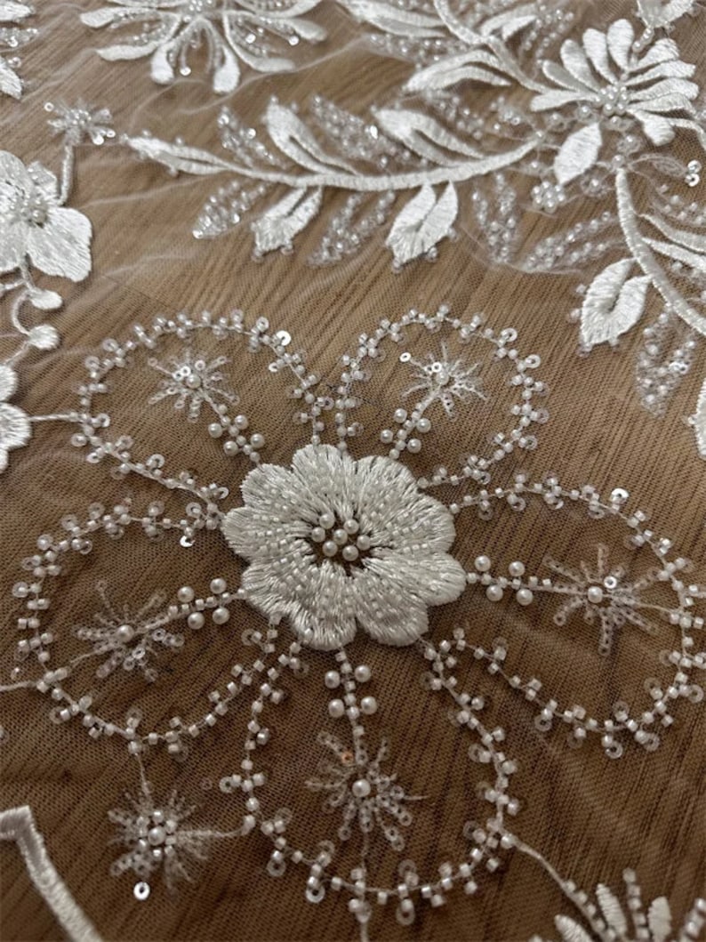 May include: White floral lace fabric with pearl and sequin embellishments. The fabric features a delicate floral pattern with intricate details. The lace is soft and flowing, making it ideal for a variety of projects.