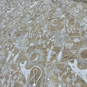 May include: A white sheer lace fabric featuring intricate embroidery and sequins. The pattern displays celestial motifs including rockets, planets, stars, suns, and crescent moons, interspersed with stylized cat figures throughout the textile.