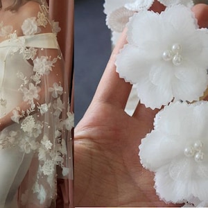 Può includere: Velo da sposa in tulle bianco con delicati ricami floreali e perline. Il velo è mostrato drappeggiato su un abito bianco.
