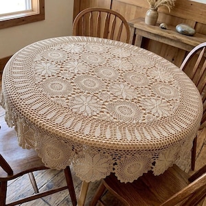 Peut inclure: Une nappe ovale en crochet beige présentant un motif répétitif de fleurs circulaires et un bord festonné. Le travail d'aiguille complexe affiche un design ajouré délicat qui recouvre la surface d'une table en bois.