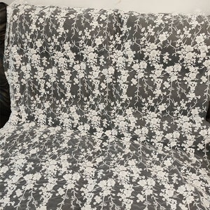 May include: White floral lace fabric. The delicate lace features a repeating pattern of white flowers and branches. The fabric is draped over a dark surface, showcasing the intricate details of the design. Ideal for bridal wear or decorative projects.