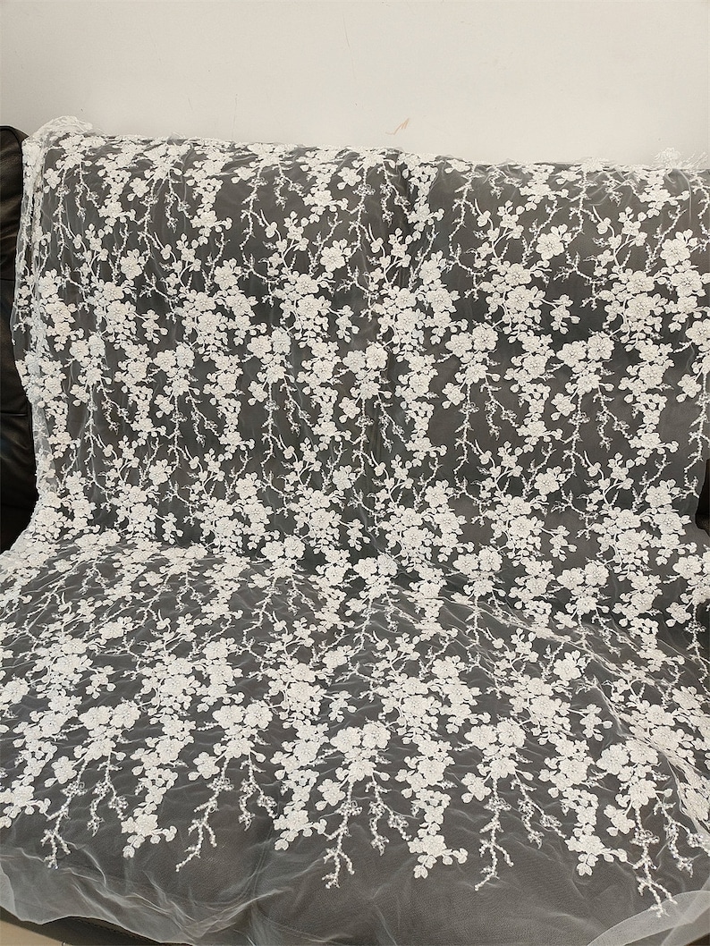May include: White floral lace fabric. The delicate lace features a repeating pattern of white flowers and branches. The fabric is draped over a dark surface, showcasing the intricate details of the design. Ideal for bridal wear or decorative projects.