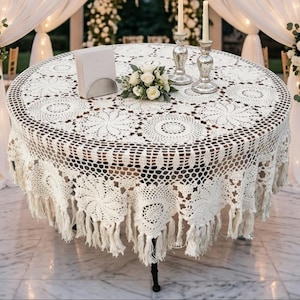 Könnte beinhalten: Eine runde, cremeweiße Häkeltischdecke mit komplizierten Blumenmustern und einem Fransenrand. Die Tischdecke liegt auf einem runden Tisch, auf dem ein kleiner Strauß weißer Rosen, zwei silberne Kerzenhalter und ein weißer Kartenhalter stehen.