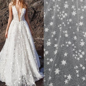 May include: A white tulle wedding dress with a plunging neckline and a star pattern. The dress is flowing and has a full skirt.