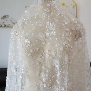 Puede incluir: Tela de novia blanca con adornos florales y lentejuelas. El material transparente está drapeado sobre un maniquí, mostrando el delicado diseño. La tela presenta pequeñas lentejuelas blancas en forma de flor y cuentas dispersas.