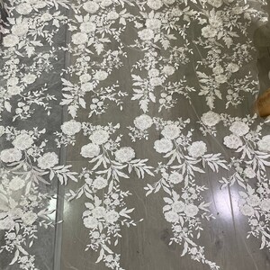 May include: White floral lace fabric with a scalloped edge. The lace features a delicate floral pattern with small white flowers and leaves on a sheer white mesh background.