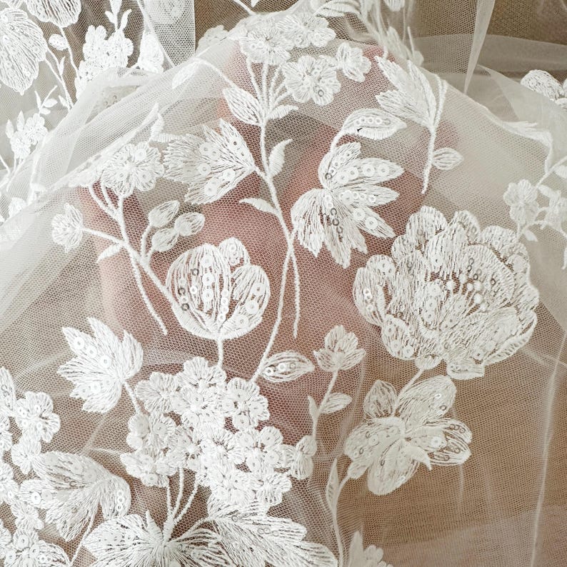 May include: White floral lace fabric with sequins. The fabric is sheer and has a delicate floral pattern. The flowers are embroidered with sequins and have a raised texture.