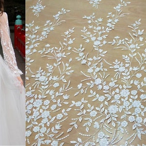 Puede incluir: Tela de encaje floral blanca con un delicado patrón de flores y hojas. El encaje es transparente y tiene una textura suave y fluida.