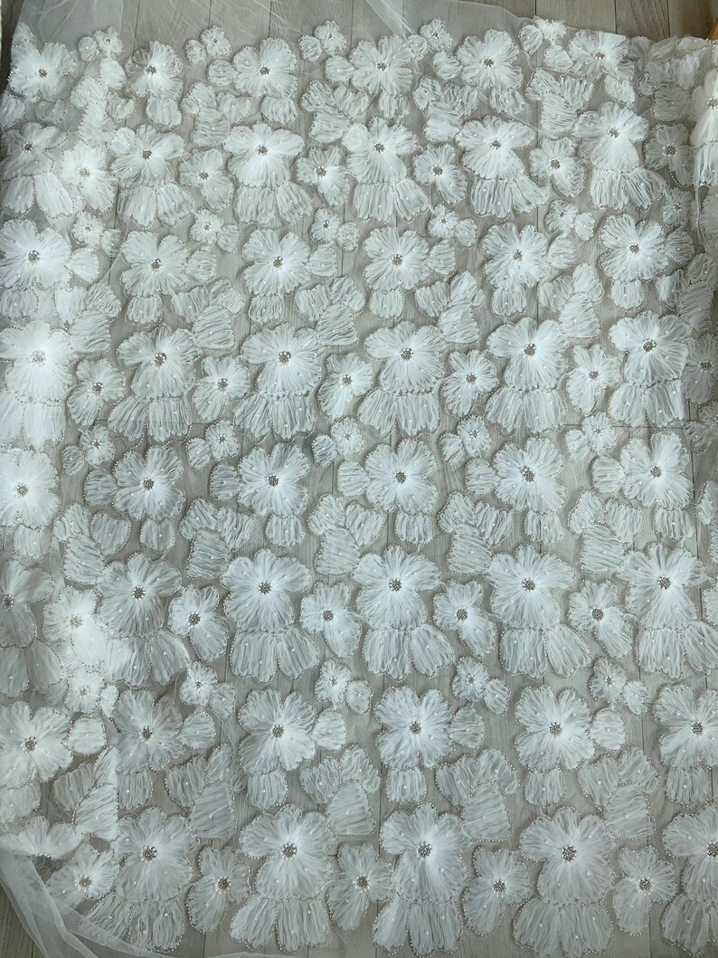 May include: White floral lace fabric with a delicate, 3D flower design. The fabric is sheer and has a soft, flowing texture.