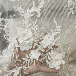 May include: White floral lace fabric with raised 3D flowers. The lace has a delicate pattern and is perfect for wedding dresses, bridal accessories, and other special occasion garments.