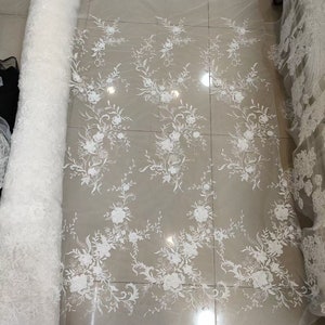 May include: White floral lace fabric with a delicate pattern of flowers and leaves. The lace is on a white background and is rolled up for display.