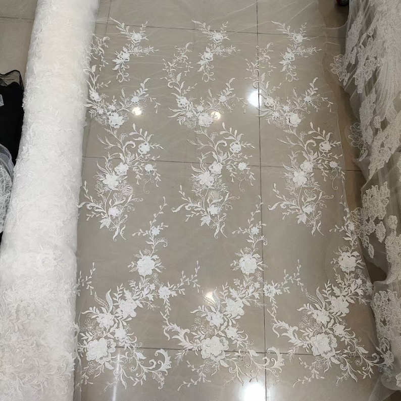 May include: White floral lace fabric with a delicate pattern of flowers and leaves. The lace is on a white background and is rolled up for display.